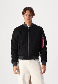 Giacca bomber in suede nera con colletto grigio, polsini e orlo a coste. Presenta una tasca laterale con zip e un'etichetta rossa. Indossata sopra pantaloni bianchi.