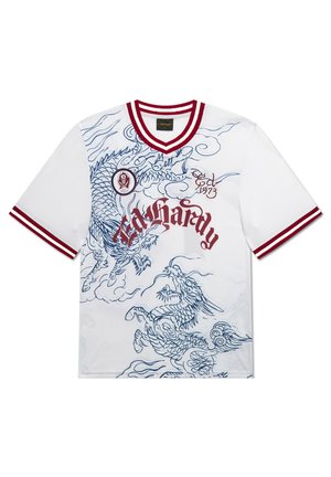 Junger Mann mit Tattoos, der ein weißes Grafik-T-Shirt, schwarze gemusterte Shorts mit Schriftzug, weiße Socken und schwarze Sneaker trägt, vor grauem Hintergrund steht.