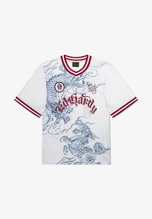 Junger Mann mit Tattoos, der ein weißes Grafik-T-Shirt, schwarze gemusterte Shorts mit Schriftzug, weiße Socken und schwarze Sneaker trägt, vor grauem Hintergrund steht.