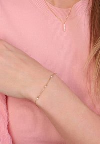 Gouden kettingarmband met kleine ronde gouden kralen en witte kralen, gedragen om een pols tegen een achtergrond van een roze geribbeld trui.