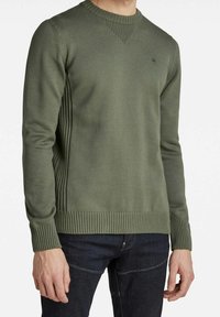 Grüner gestrickter Pullover mit geripptem Ausschnitt und Saum, seitlichen vertikalen Rippenstrukturen und einem kleinen markanten Akzent auf der Brust.