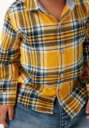 Barn iført en gul, sort og hvid ternet skjorte med knapper og et lille broderet mærke samt blå jeans, hænderne hvilende ved siden.