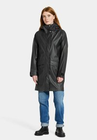 Svart vattentät regnjacka med hög krage, tryckknappar och två framfickor; i kombination med blå jeans och svarta ankelkängor.