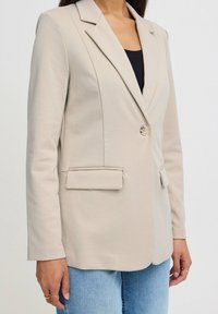Světlý béžový tailored blazer s jedním knoflíkem, klopami, dvěma předními kapsami a hladkou strukturou. Nesený přes černý top.