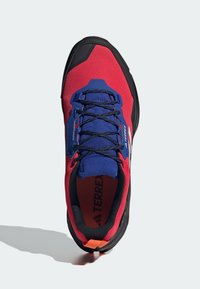 Punainen ja sininen verkkokankaasta valmistettu urheilukenkä mustilla yksityiskohdilla, pyöreä kärki, teksturoitu yläosa ja oranssi yksityiskohta kannassa. Nauhat ovat tummansiniset.