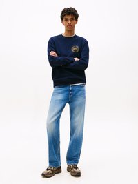 Felpa navy con logo circolare dorato, vestibilità comoda. Jeans light blue a gamba dritta con design sbiadito. Sneakers marroni, con texture.