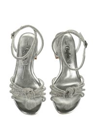 Ezüst, pántos magassarkú szandál, fémes fényzéssel, díszes csomó részlettel és bokapántokkal. Klasszikus nyitott orrú kialakítás.
