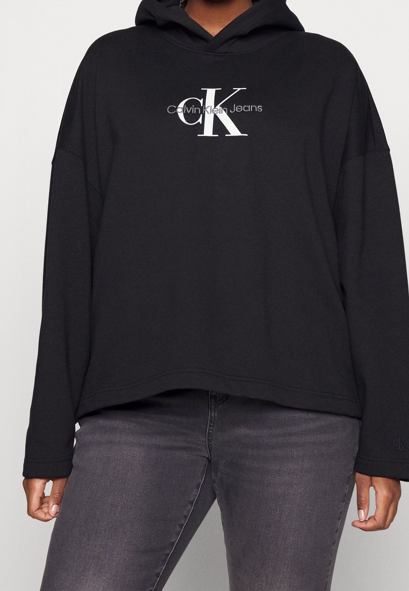 Personne portant un sweat à capuche noir Calvin Klein Jeans avec un grand logo blanc "cK" et un jean gris, visible du cou aux hanches.