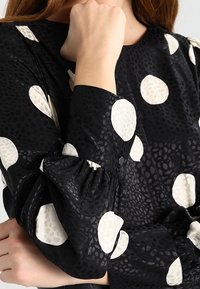 Blouse noire à motifs avec de grands pois crème, tissu texturé, manches froncées et détail de bouton au niveau du poignet.