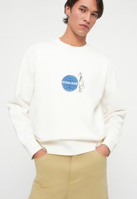 Młody mężczyzna z ciemnymi włosami w białej bluzie z kapturem, na której znajduje się niebieska kula ziemska z napisem "PAN AM" i małym rysunkiem mężczyzny trzymającego teczkę.