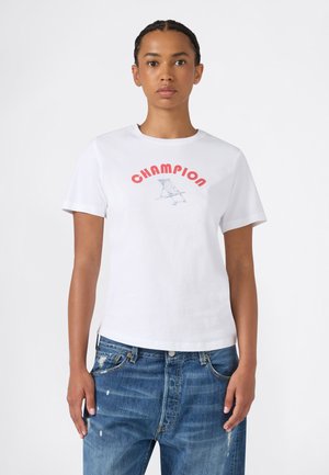 Femme portant un T-shirt blanc avec un texte rouge "CHAMPION" et un dessin de chaise de plage bleue, associé à un jean bleu déchiré.