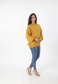 Maglione oversize senape con colletto a coste e maniche a palloncino, abbinato a jeans blu slim-fit e sandali con tacco trasparente.