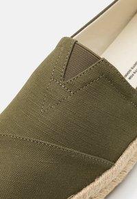 Zelene platnene cipele bez vezivanja s teksturiranim gornjim dijelom i prošivenim detaljima, s potplatom od jute koji pruža dodatno prianjanje i stil.
