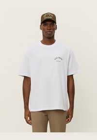 Witte T-shirt met korte mouwen, voorzien van een klein zwart logo op de borst. Gekozen in combinatie met een groene pet met de merknaam in witte tekst.