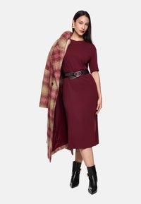 Bordeauxrotes Kleid mit kurzen Ärmeln und seitlicher Schlitze, kombiniert mit einem karierten langen Mantel. Schwarze Knöchelstiefel mit goldenen Akzenten runden den Look ab.