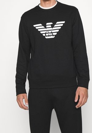 Mężczyzna ubrany w czarną bluzę z długim rękawem z białym logo orła w paski oraz literami "GA" na piersi.