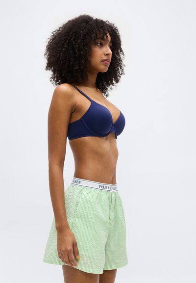 Γυναίκα με σγουρά μαλλιά που φοράει μπλε σουτιέν και ανοιχτό πράσινο σορτς Polo Ralph Lauren, στέκεται πλευρικά πάνω σε μια απλή φόντο.