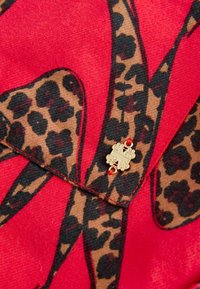 Punane kangas, millel on leopardimustriga disain, kaunistatud kuldse embleemi ja punase vääriskiviga. Sile tekstuur koos kontrastsete mustritega.