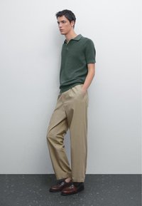 Polo shirt verde scuro a maniche corte, pantaloni beige a pieghe e scarpe marroni con lacci, su uno sfondo grigio chiaro.