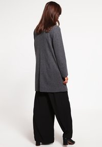 Cappotto lungo in misto lana grigio con taglio dritto, tasche laterali e colletto a risvolto, indossato sopra pantaloni neri larghi. Design semplice e pulito.