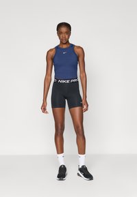 Ναυτικό αμάνικο μπλουζάκι με λογότυπο Nike, συνδυασμένο με μαύρα σορτς Nike Pro που διαθέτουν λευκή ζώνη. Το μοντέλο στέκεται σε ουδέτερο περιβάλλον.