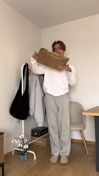 Egy fehér, hosszú ujjú pólót viselő személy egy barna csomagot tart. Szürke melegítőnadrágot és bézs, texturált felületű papucsot visel.