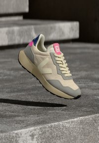 Μπεζ αθλητικό παπούτσι με υφασμάτινο άνω μέρος με υφή, γκρι σουέντ λεπτομέρειες, ροζ και μπλε τμήματα στη φτέρνα, και μαύρη λαστιχένια εξωτερική σόλα.