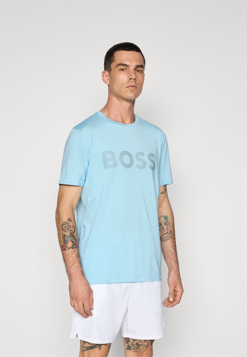 T-shirt bleu clair à manches courtes avec le mot "BOSS" en gris foncé sur la poitrine. Tissu doux avec une coupe décontractée, associé à un short blanc.