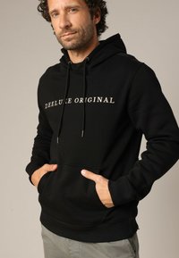 Zwarte hoodie van zacht materiaal met een voorste kangoeroezak en de tekst "DEELUKE ORIGINAL" in metallic goud. Koordjes aan de capuchon.