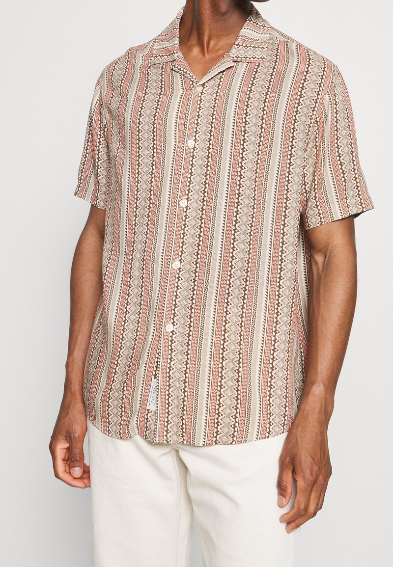 Homme portant une chemise à manches courtes à boutons avec des motifs tribaux verticaux rouges, bruns et beiges, assortie à un pantalon clair.
