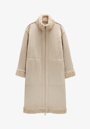 Beżowy płaszcz z wysokim kołnierzem i zamkiem błyskawicznym z przodu. Miękkie, teksturowane akcenty z baraniej wełny na kołnierzu i rękawach. Dwie duże kieszenie z przodu.