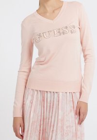 Różowy sweter z długim rękawem i dekoltem w serek, ozdobiony napisem "GUESS" z kryształków na piersi, zestawiony z plisowaną spódnicą w kwiatowy wzór.