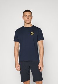 Temno modra majica s kratkimi rokavi, kroja crew neck, ki ima majhen rumen geometrijski logotip na levem prsnem delu. Narejena iz mehkega, lahkega materiala.