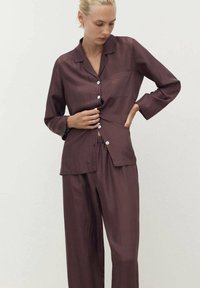 Set di pigiama in satin bordeaux composto da una camicia con bottoni dotata di taschino sul petto e maniche lunghe, abbinata a pantaloni con vita elastica.