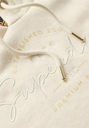 Tissu de hoodie beige clair avec un logo "Superdry" brodé en blanc et or, doté de cordons blancs avec des embouts en métal doré.