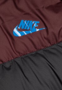 Nike tuulenpitävä takki, jossa on maroon yläosa ja sininen logo sekä musta alaosa. Luja rakenne ja näkyvät ompeleet.