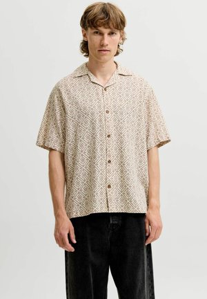 Jeune homme aux cheveux châtain clair portant une chemise beige à manches courtes avec un motif géométrique en losanges et un pantalon noir.