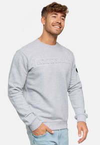 Sudadera gris clara con texto en relieve en el frente, puños y dobladillo acanalados, y un cuello redondo. La tela parece suave y texturizada.