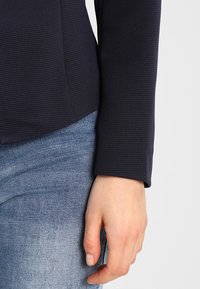 Handen på en person som bär en mörk, ribbad långärmad tröja och blå denimjeans, vilar vid höften mot en neutral bakgrund.