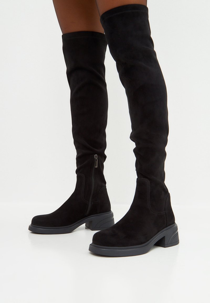 Botas negras de ante sobre la rodilla, con cremallera lateral, tacón cuadrado y textura suave. Diseñadas con una silueta ajustada.