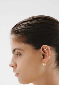 Pendientes en tono dorado con un diseño de concha y un aro curvado. La modelo tiene un peinado elegante, lo que resalta claramente la joyería contra un fondo claro.