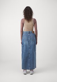 Denim maksi svārki ar taisnu piegriezumu, divām sānu kabatām un aizmugures kabatām, vidēji zilā krāsā. Apvienoti ar béžīgu bezpiedurkņu topu un sporta apaviem.