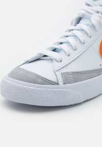 Vit sneaker med grå mockatoe, texturerad sula, vita snören och partiell orange detalj på sidan.