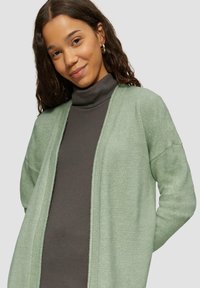 Cardigan vert clair avec une finition texturée, à l'avant ouvert et à manches longues, porté par-dessus un haut col roulé gris foncé. Design minimaliste.
