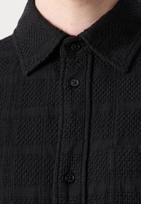 Zwarte shirt met textuur, voorzien van een geweven patroon, een kraag en zwarte knopen aan de voorkant. Close-up weergave die zich richt op de materiaaleigenschappen en ontwerpdetails.