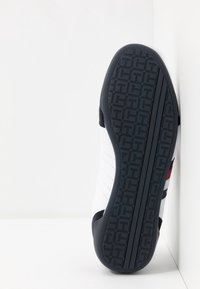 Vit sneaker med svart sula, försedd med geometriskt mönster på sulan, vilande på en vit yta mot en vit bakgrund.