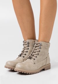 Bottines à lacets beige avec semelles en caoutchouc épaisses portées sur des jambes nues sur fond blanc.