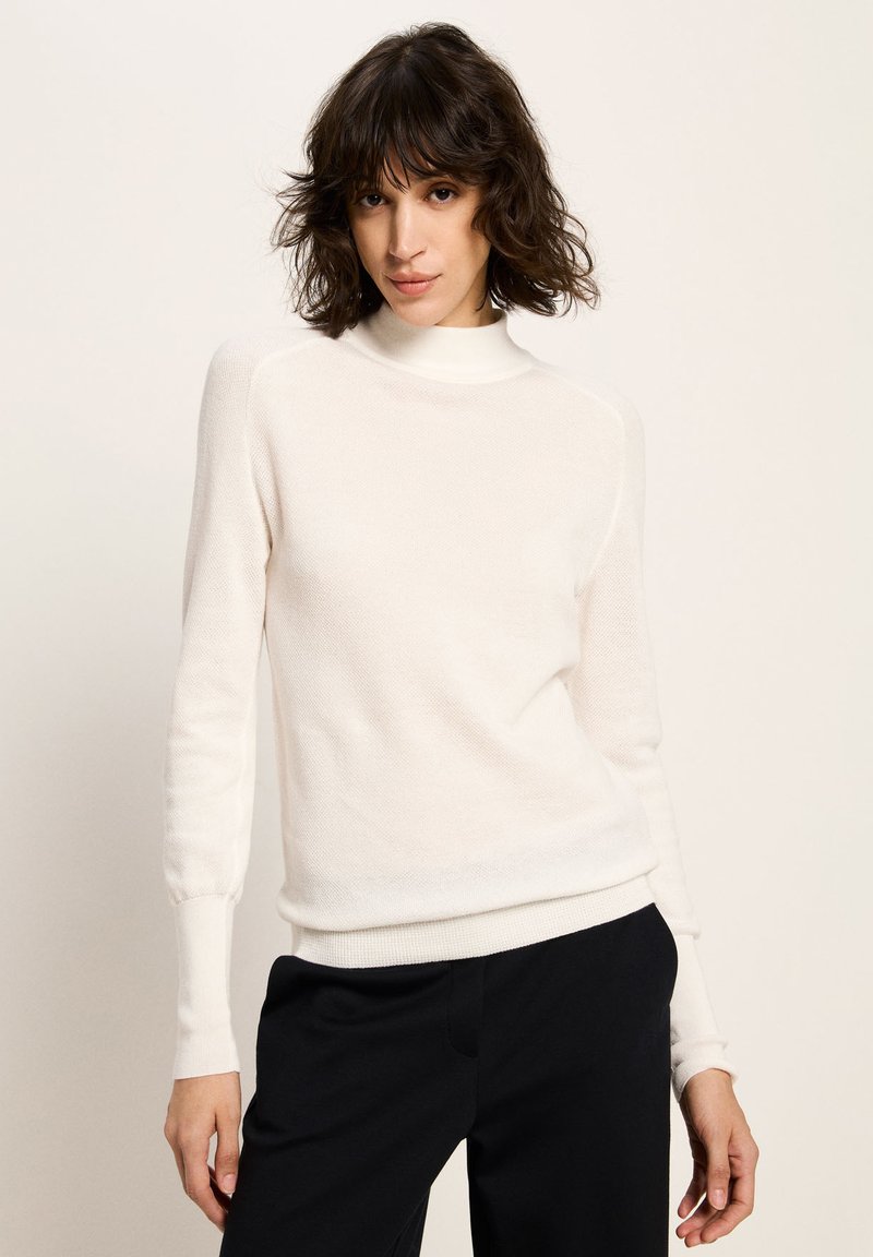 Eine Frau mit kurzen, welligen, dunklen Haaren trägt einen weißen Langarm-Pullover mit Rollkragen und schwarze Hosen vor einem schlichten, hellen Hintergrund.