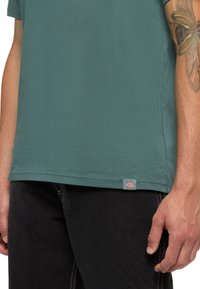 Zelené bavlněné tričko s kulatým výstřihem a krátkým rukávem, vybavené malou logem zašitým na spodním lemu. Nosené s černými džíny.