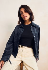 Veste-chemise en daim bleu foncé avec détails à franges, sous-pull col rond côtelé noir, associée à un pantalon beige et une ceinture argentée.